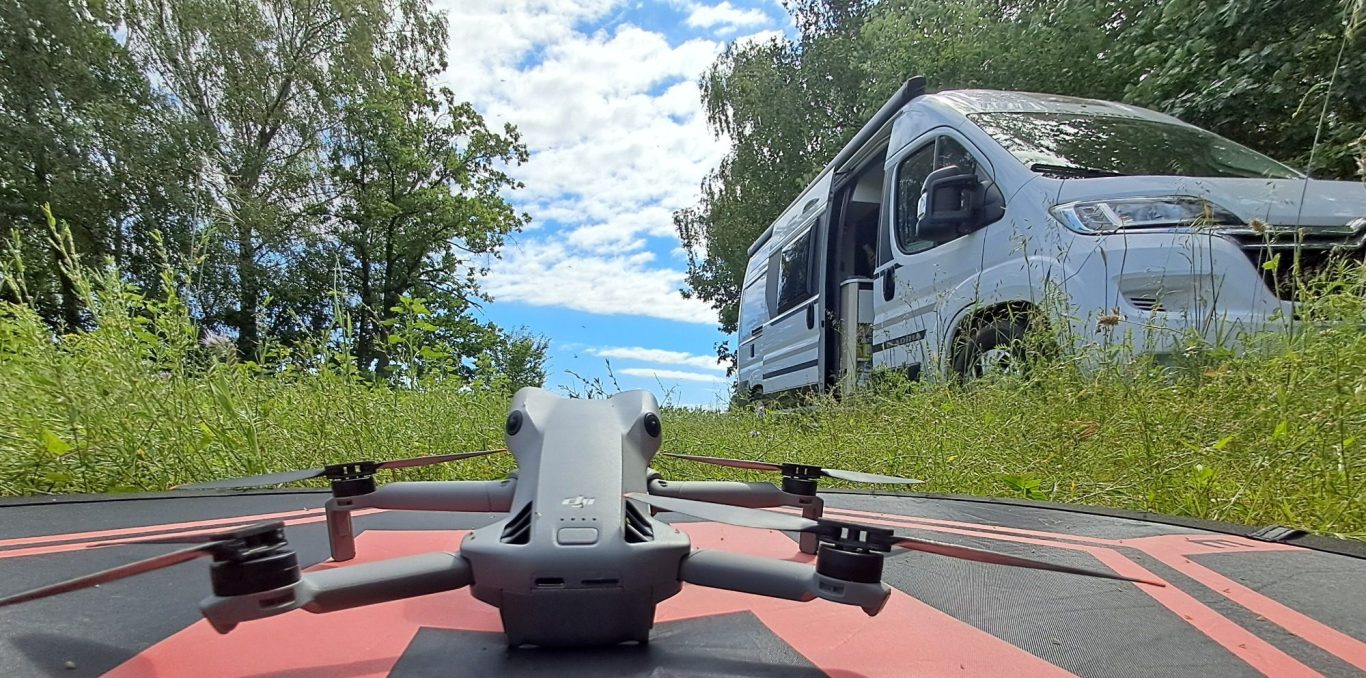 Drohne im Vordergrund, Wohnmobil im Hintergrund, umgeben von grüner Landschaft und blauem Himmel.